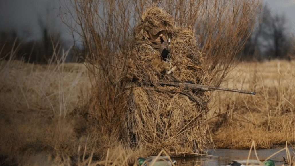 A waterfowl hunter in a camouflage suit standing along a river holding a rifle