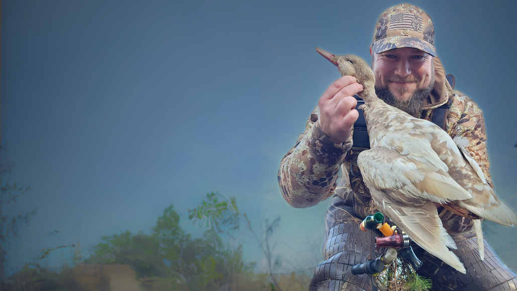 A waterfowl hunter in camouflage holding a duck in the air.