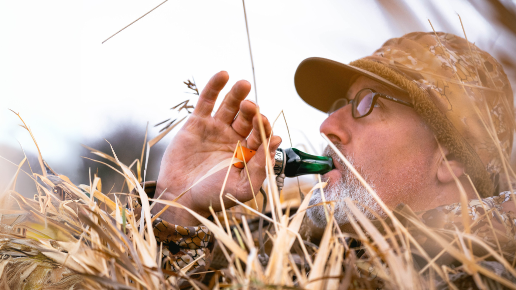 waterfowl hunter dressed in camo in a layout blind blowing Slayer's DUBAR duck call