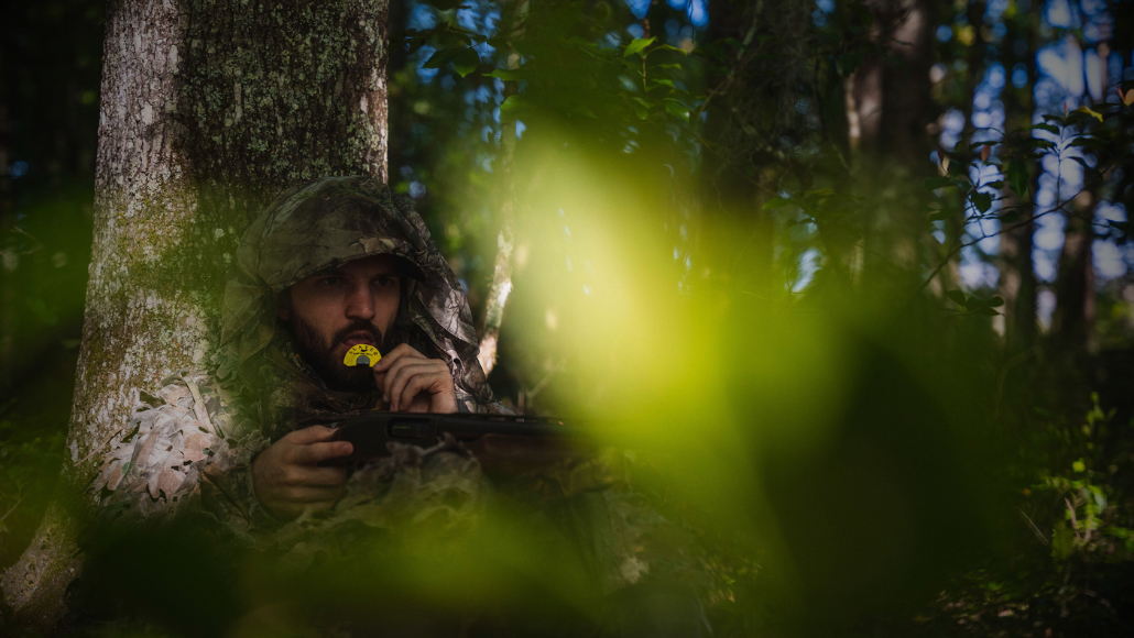 A turkey hunter in camouflage is sitting in the woods using a turkey mouth call