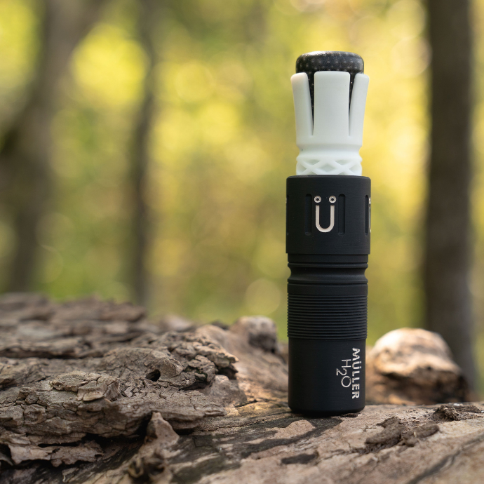 Black and white single reed ATC - Muller Chokes collaboration duck call with the Muller logo and H2O branding on a log with a blurred wooded background.