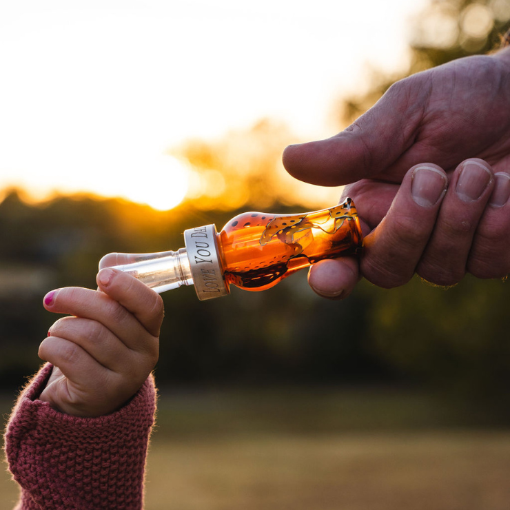 Child’s hand reaches for a custom engraved duck call personalized with text “LOVE YOU DAD,” passed down from an adult at sunset — symbolizing hunting tradition and family legacy.