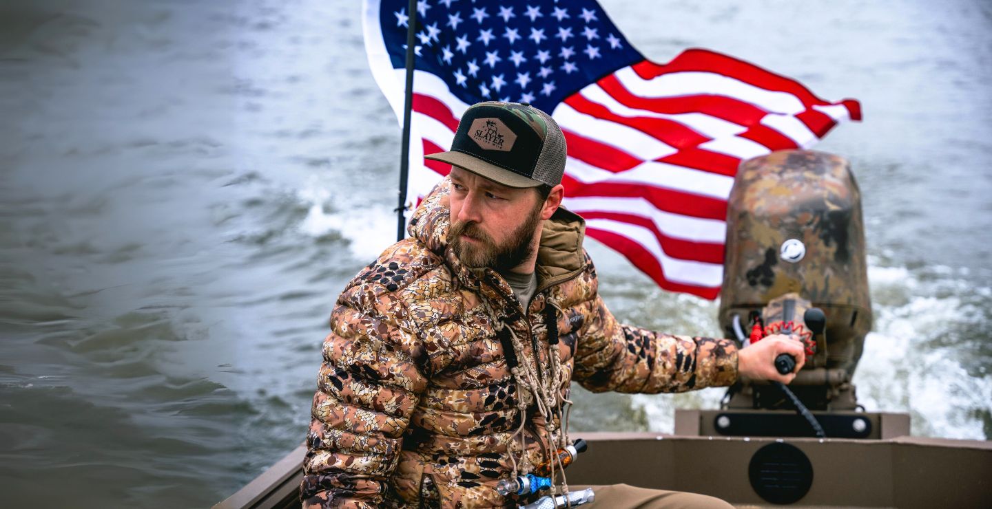 Waterfowl hunter in camouflage steering a boat with an American flag flying behind, showing patriotism and passion for the hunt.