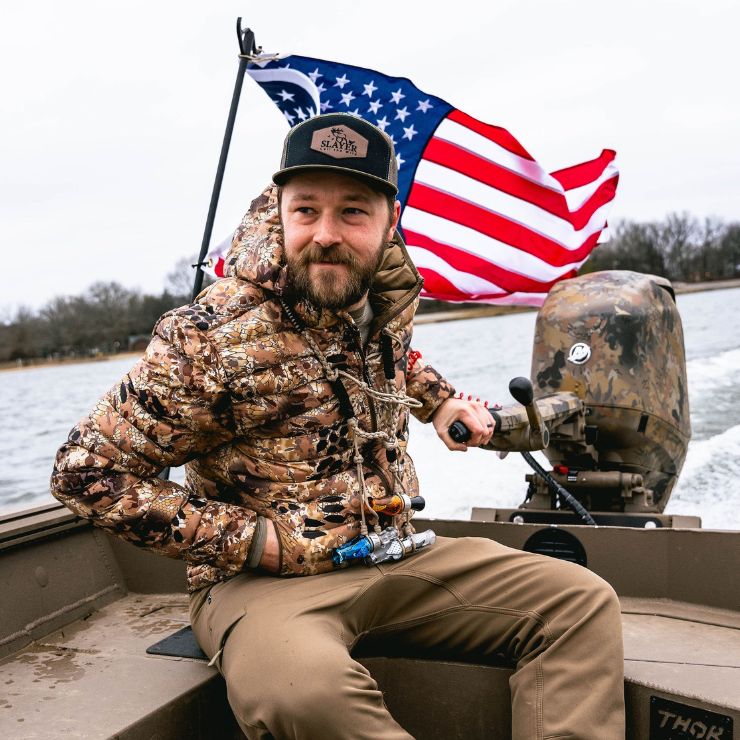 Waterfowl hunter in camouflage steering a boat with an American flag flying behind, showing patriotism and passion for the hunt.