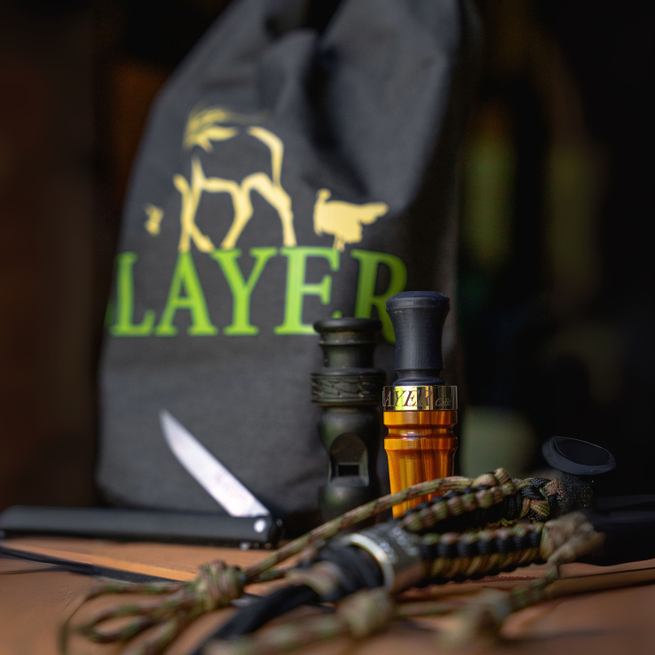 A dark gray Slayer dry bag with a bright green logo and elk silhouette, sitting atop a tan YETI cooler. In front of the bag are two duck calls, a field dresser, and a braided waterfowl lanyard.