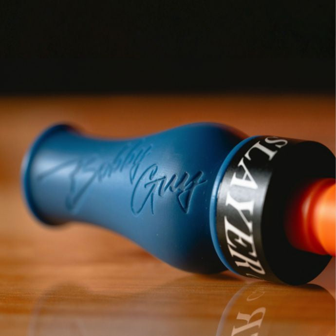 A close-up of the blue and orange Bobby Guy Signature Series duck call with an embossed script signature, a black band reading 'SLAYER' in white, resting on a wooden surface.