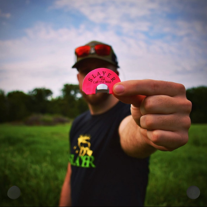 Hunter holding a Slayer diaphragm elk call labeled SLAYER with a blurred grassy field and trees in the background, wearing a Slayer t-shirt and sunglasses.