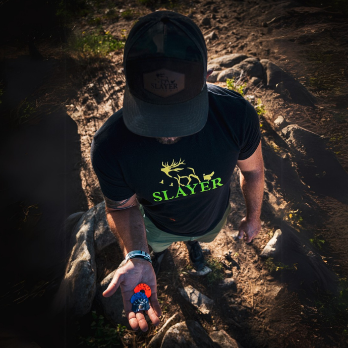 Top-down view of a hunter in a Slayer hat and shirt holding multiple diaphragm elk calls in his open palm while standing on a dirt trail surrounded by rocks and sunlight.