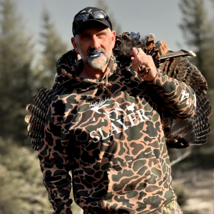Slayer camo hoodie worn by a turkey hunter in the field, featuring a bold white logo and front pouch pocket — rugged, comfortable, and built for the hunt.