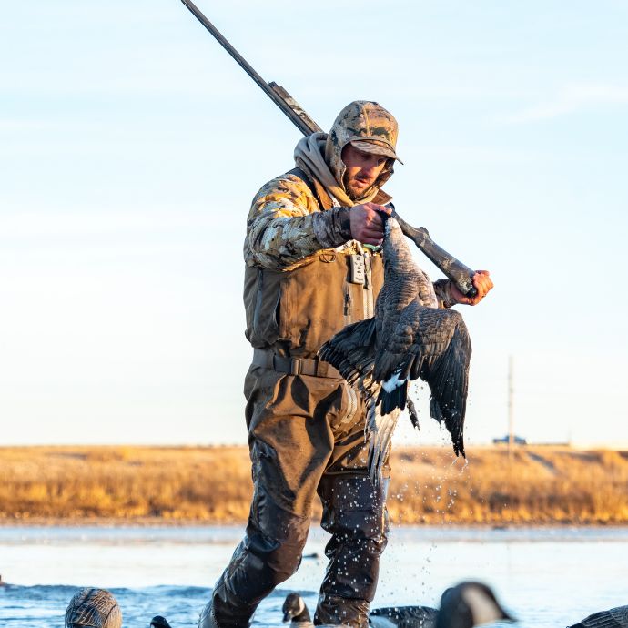 A duck hunter in camouflage clothing stands in shallow water holding a duck with a shotgun, with a marshland landscape and pale sky in the background.
