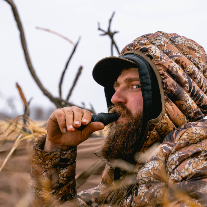 A bearded hunter in a camouflage jacket and hood uses a duck call while concealed in tall grass. He is intently focused, likely watching for approaching waterfowl, with leafless trees and overcast sky in the background.