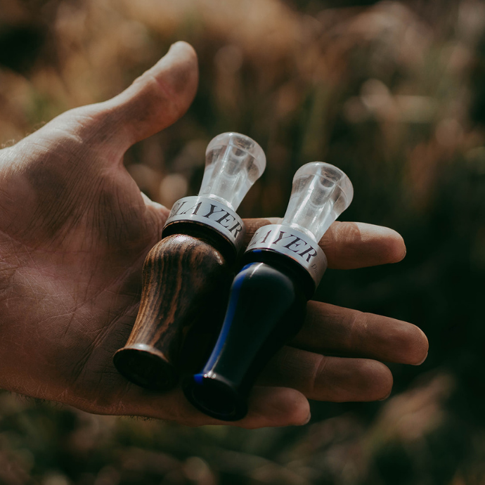 A hunter holds two duck calls in one hand: the Suzie Slayer in rich wood grain and the Drake Slayer in Black Triple Curle acrylic with a thin blue line, honoring law enforcement. Both calls feature clear tone boards and silver bands engraved with "SLAYER," set against a blurred natural background.