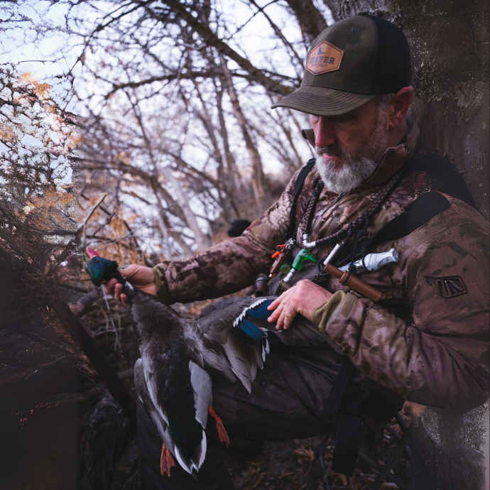 A bearded hunter in camo clothing and a Slayer Calls hat examines a harvested duck while sitting against a tree in a late-season, leafless forest. Multiple duck calls hang from lanyards around his neck, including bright green, white, and wood-tone calls — including the Suzie Slayer wood duck call. The duck’s colorful plumage is visible, contrasting with the muted tones of the wooded background.
