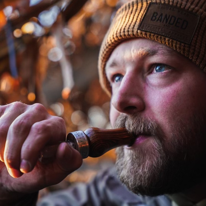 Close-up of a bearded hunter wearing a brown knit "Banded" beanie while blowing the Suzie Slayer duck call in natural wood. The silver band engraved with "SLAYER" is visible, and warm morning light filters through the camo blind in the background.