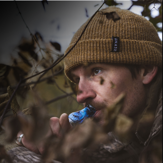 Waterfowl hunter concealed in thick brush uses the Slayer DUBAR double reed duck call in Blue Wing Mesh — designed for reliable performance and realistic sound in any hunting conditions.
