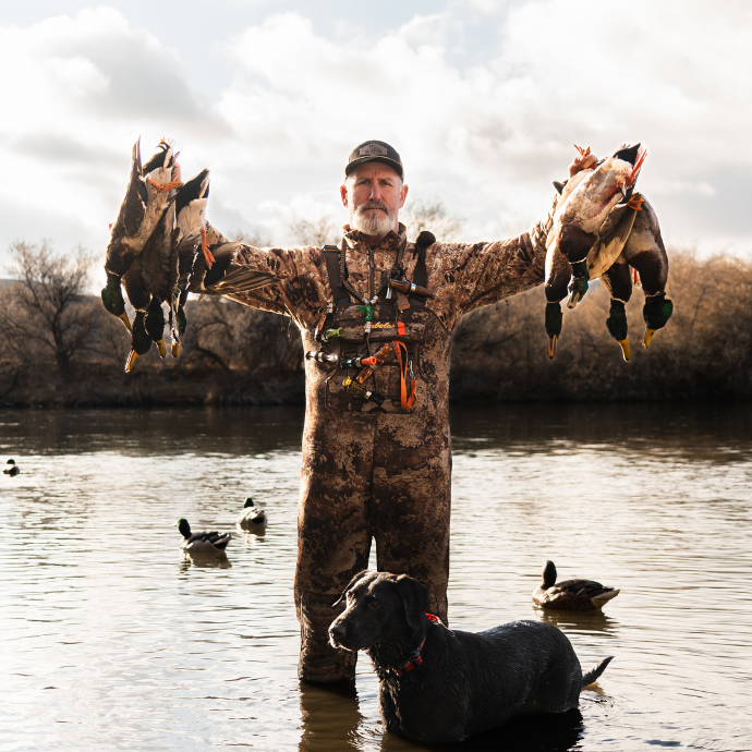 Hunter standing knee-deep in a river holds up several mallards with a black retriever beside him. Decoys float on the water, with late-season brush and trees in the background.