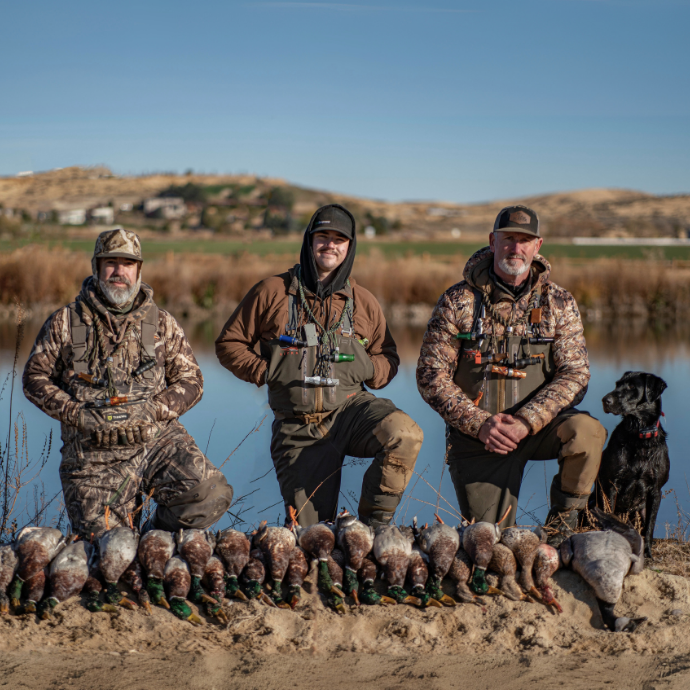 Three camo-clad hunters kneel by a pond with a long line of harvested mallards in front of them. A black Labrador retriever sits at their side, and autumn hills rise in the background. Men sport the DUBAR single reed duck call