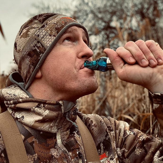 Hunter in camo uses the Slayer DUBAR single reed duck call in Blue Wing Mesh, delivering crisp, high-volume calls in marshy waterfowl habitat.