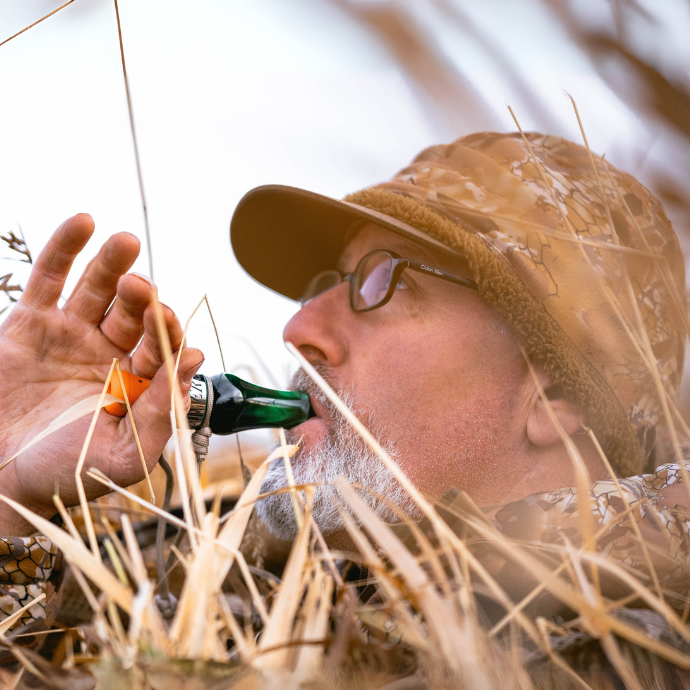 Waterfowl hunter in camo and glasses uses a green and orange DUBAR double reed duck call while concealed in tall grass, fully hidden and focused during a call sequence.