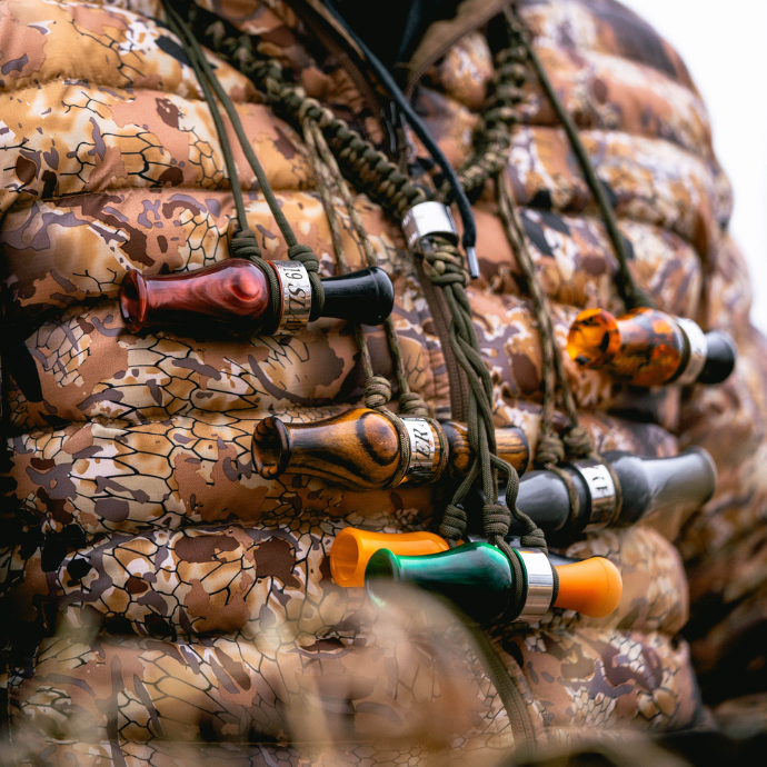 Close-up of a camo-clad waterfowl hunter wearing a paracord lanyard with multiple Slayer duck and goose calls in various colors and materials — ready for any calling scenario in the blind, featuring the DUBAR double reed duck call in green pearl with orange insert.