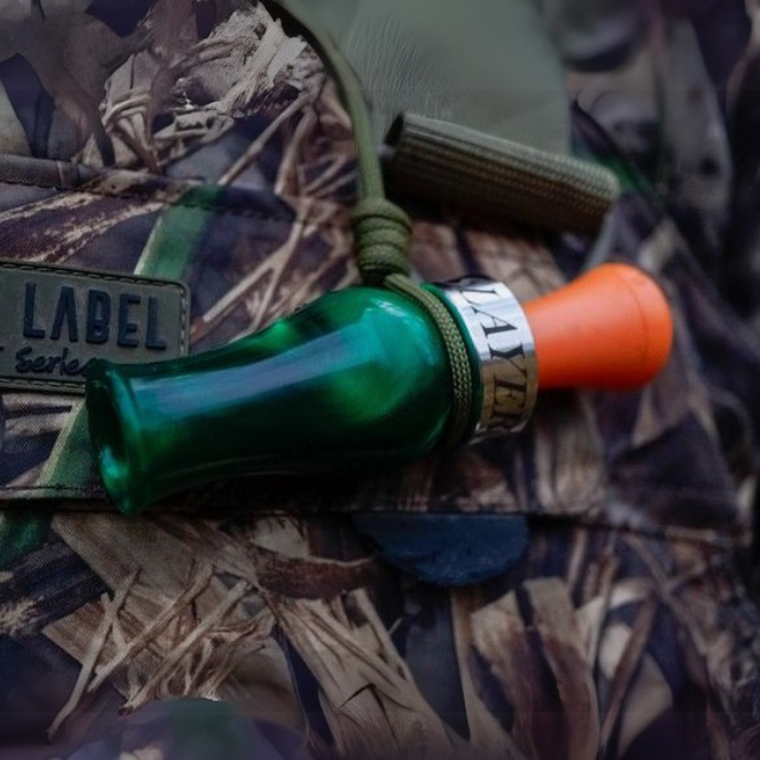 Close-up of the Slayer DUBAR double reed duck call in green pearl with an orange insert, clipped to a camo lanyard on a hunter’s waterfowl jacket.