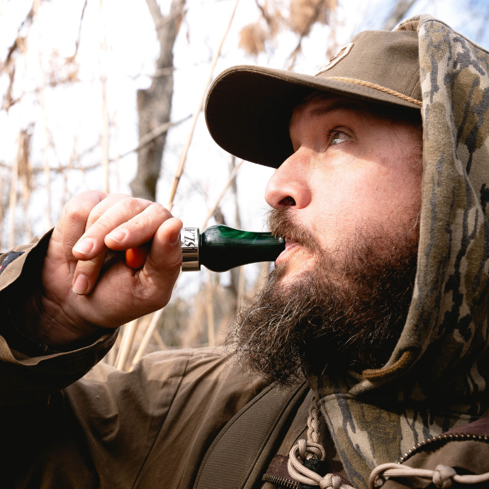 Waterfowl hunter using a DUBAR single reed duck call in Green Pearl with orange insert during a cold-weather duck hunt in the reeds.
