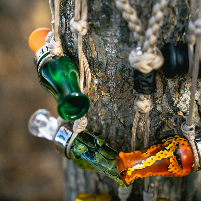 Close-up of DUBAR single reed duck call in Green Pearl and Orange, alongside the Drake Slayer duck call in Green Marsh and Citrine Water, hanging on paracord lanyards against tree bark.