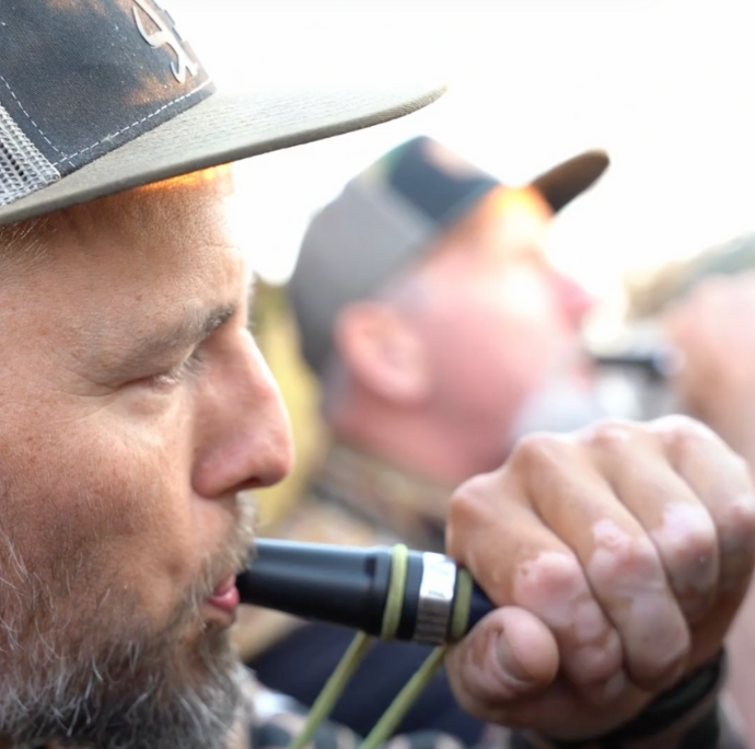 Close-up of a bearded waterfowl hunter blowing a black Slayer hybrid duck call, with fellow hunters in the background during a group calling session.