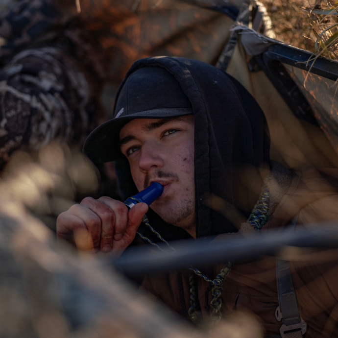 Waterfowl hunter in a layout blind using a blue Slayer hybrid duck call called the Yolo Bypass wearing a black hoodie and hat, focused during a cold-weather hunt.