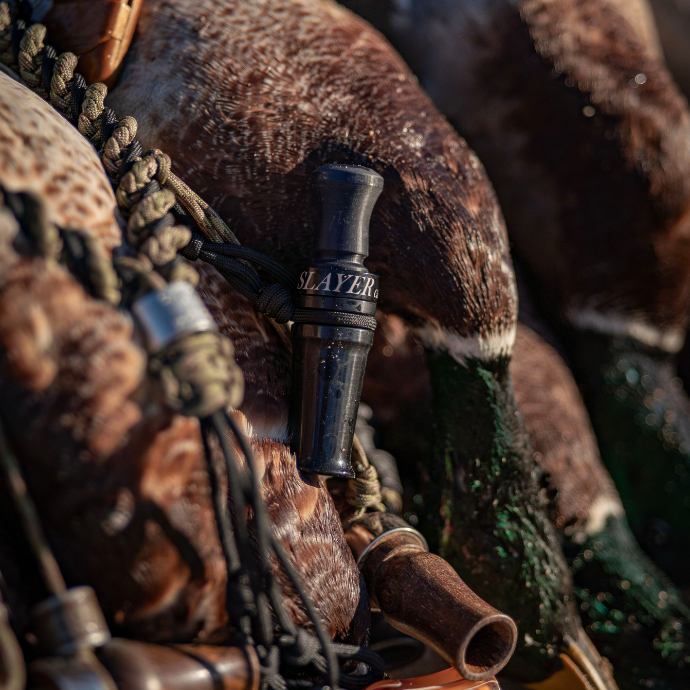 Slayer Calls' Yolo Assassin Limited Edition duck call shown in the field, hanging from a lanyard against a background of harvested mallards. This hybrid acrylic duck call features cutdown-style volume and rasp, ideal for breaking ducks from a distance in flooded timber or open water hunts.