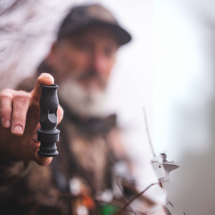 Waterfowl hunter holding up a black Slayer duck whistle, designed for drake mallard, pintail, wigeon, and teal calls — blurred figure and misty blind in background.