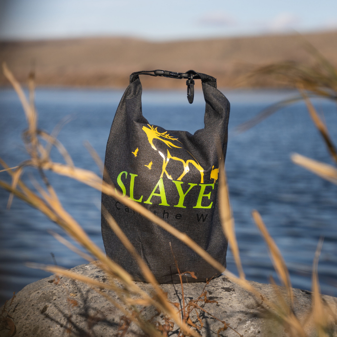 Slayer branded roll-top dry bag with neon elk logo, staged on a riverbank rock — water-resistant gear bag built for hunters and rugged outdoor use.
