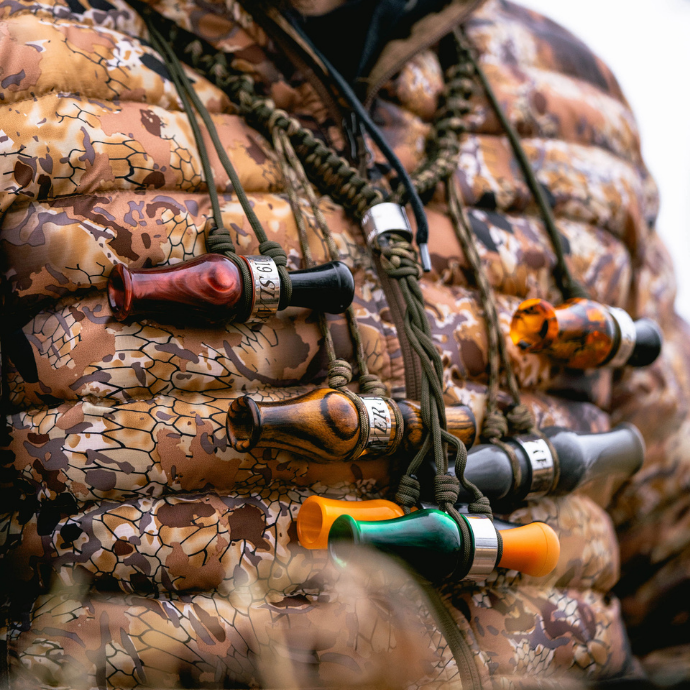 Close-up of a hunter in camo jacket wearing a lanyard with multiple Slayer duck calls in various custom acrylic finishes, ready for the field.