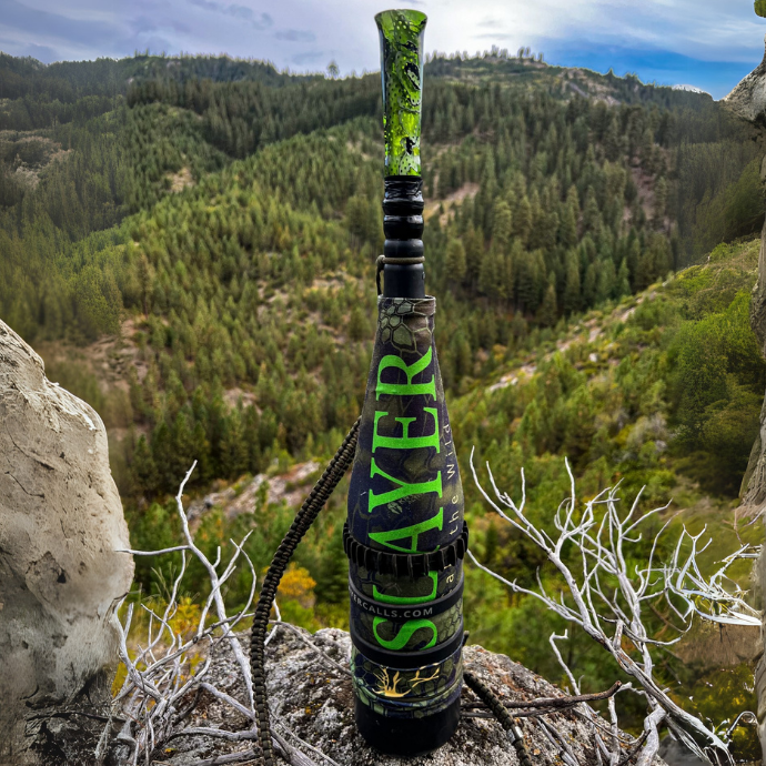 ArchAngel elk bugle is shown with Kryptek camo cover and green marsh acrylic mouthpiece placed on a boulder overlooking Idaho mountain landscape