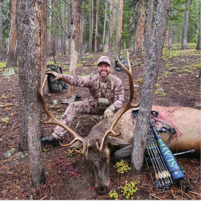 Elk hunter kneeling beside a freshly harvested mature bull elk after a successful hunt using the Enchantress push button cow call and attachable bugle tube by Slayer Calls in the Idaho wilderness.