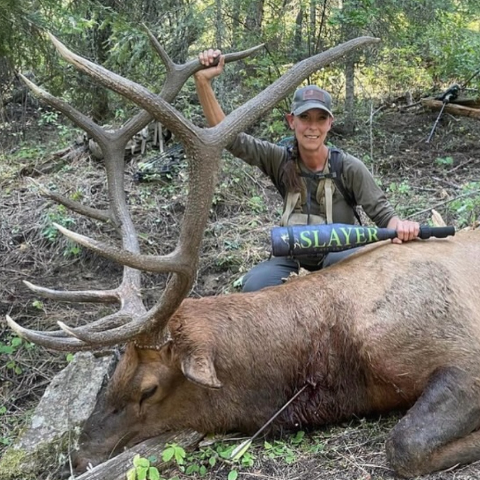A female elk hunter proudly poses beside a large 5x6 mature bull elk after a successful hunt, having used Slayer Calls' Enchantress elk call combo to call in the animal.