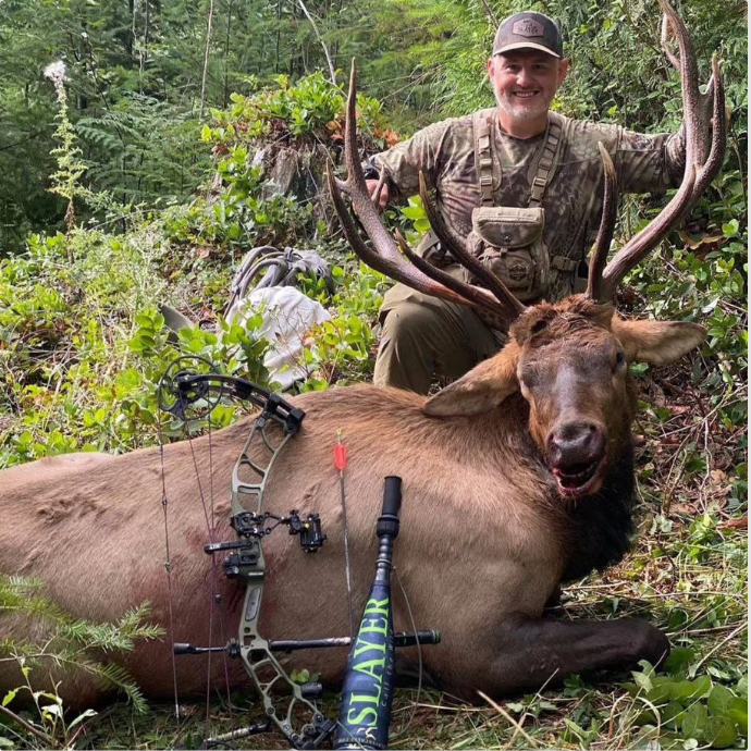 Archery elk hunter posing with a Roosevelt elk in the Oregon wilderness, showcasing the mature bull called in using the Enchantress cow call -bugle combo by Slayer Calls.