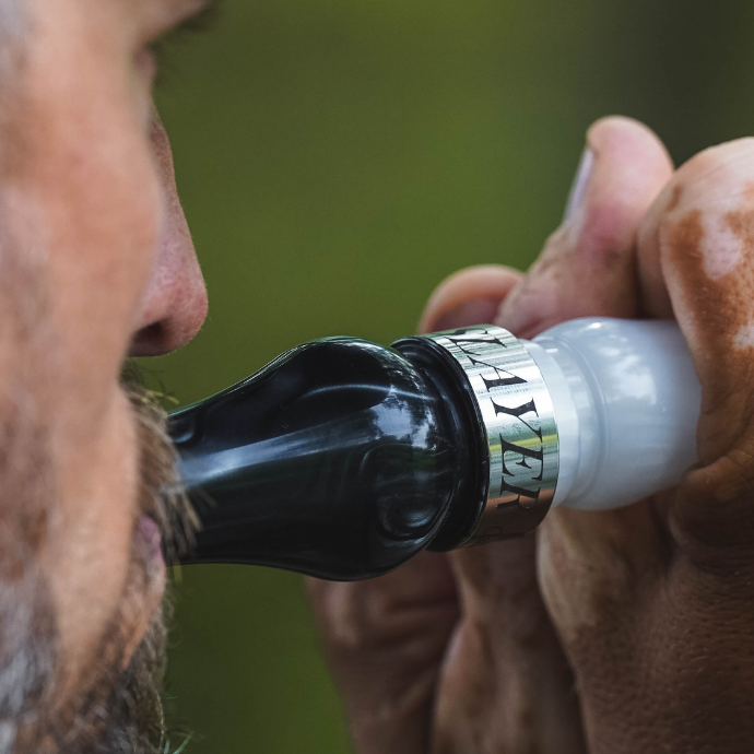 Close-up of a hunter blowing the Honker Slayer goose call in black and white pearl acrylic. The silver band engraved with "SLAYER" is clearly visible, and the background is softly blurred with natural green tones.