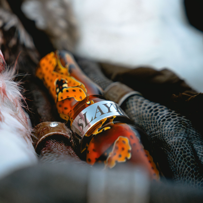 Close-up of the Honker Slayer goose call in vibrant citrine orange acrylic with black accents. The call rests among feathers and textured hunting gear, with the word "SLAYER" engraved on the metal band.