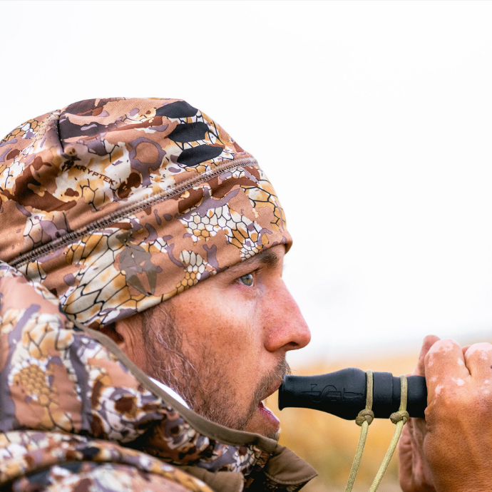Waterfowl hunter in Kryptek camo using a black Slayer Dagger goose call in the field — focused and calling during a cold-weather Canada goose hunt.