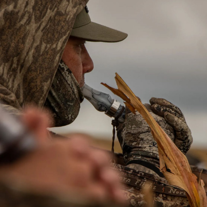 A camouflaged hunter uses the Honker Slayer goose call in snow mesh acrylic while concealed in a field blind. The hunter grips the call with gloved hands, blending into the natural surroundings of dry corn stalks under an overcast sky.