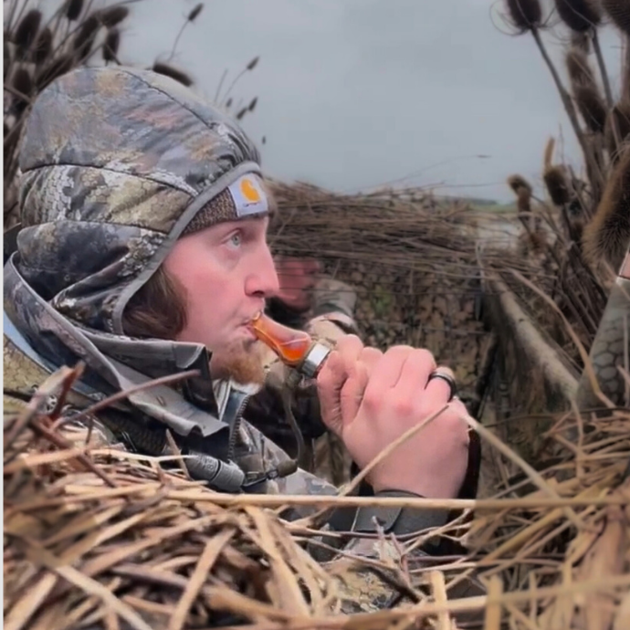 Waterfowl hunter in a camo blind using the Tar Belly specklebelly goose call from Slayer Calls, wearing cold-weather gear and surrounded by natural brush cover.