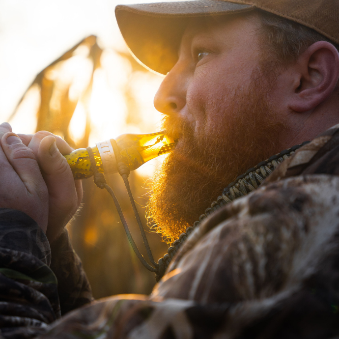 Hunter using Slayer’s Tar Belly specklebelly goose call in yellow acrylic, calling at sunrise while wearing camouflage gear and a lanyard full of calls.