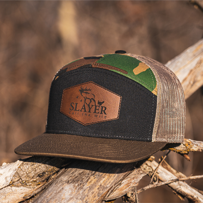 Slayer Calls camo trucker hat with leather patch logo — mesh-back design with green camo and flat bill, built for hunters who live the Call the Wild lifestyle.