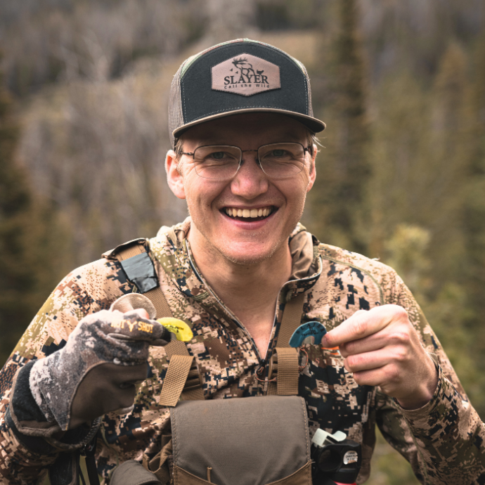 Hunter smiling in camo with elk reeds in hand, wearing the Slayer trucker hat in camo — breathable mesh-back cap built for comfort and durability in the backcountry.