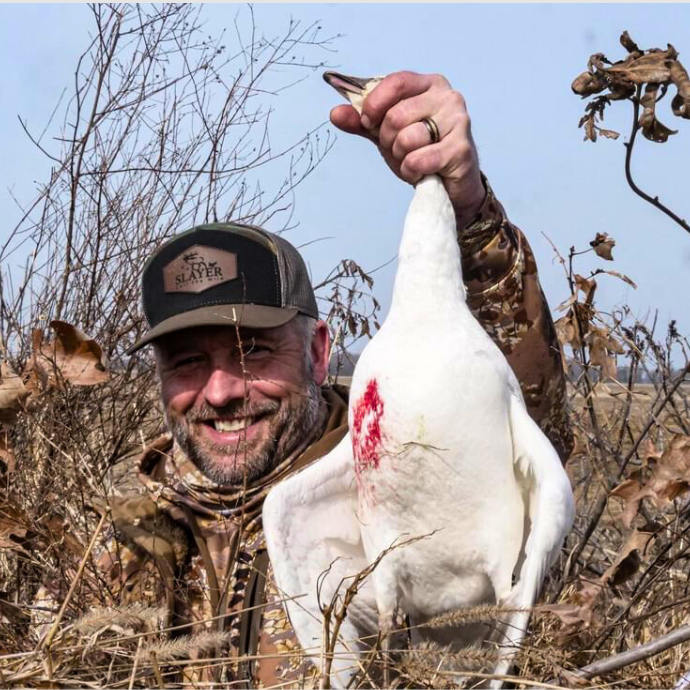 Smiling waterfowl hunter in camo holding a harvested snow goose, wearing a Slayer Calls camo patch hat — durable outdoor Slayer trucker hat available in camo.