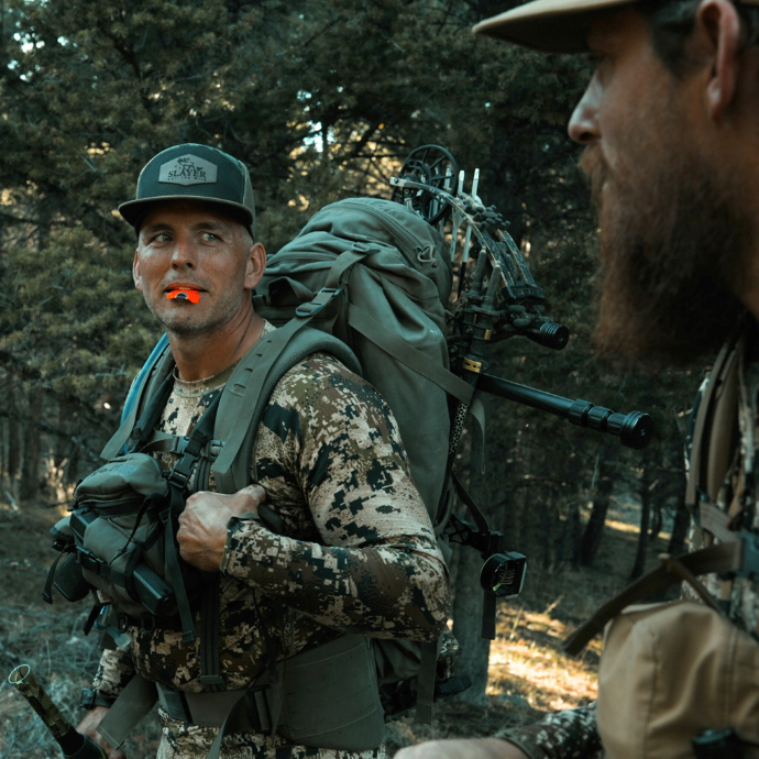 Pair of elk hunters dressed in camo hiking through the woods. One of the hunters is using Slayer's red Miner elk reed from the Clearwater Series.