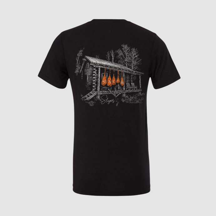 Black t-shirt with a graphic design of orange game bags hanging from the eves of an old hunting cabin on the back against a light gray background