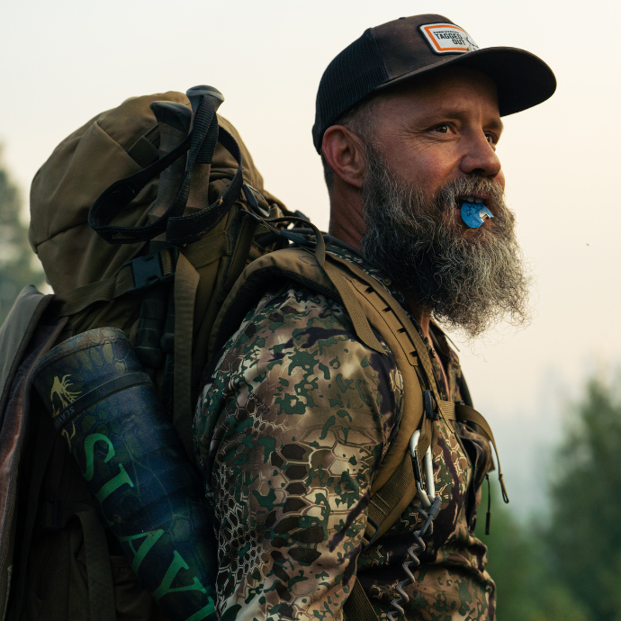 Elk hunter wearing camouflage uses Slayer Calls’ blue Selway elk reed; Archangel bugle tube secured in hunting pack.
