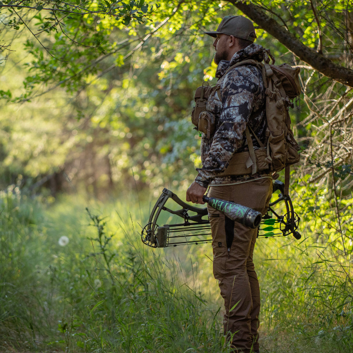 Elk hunter in camouflage gear with a backpack, bow and Slayer's LIl' Warrior bugle tube walking through a forest.